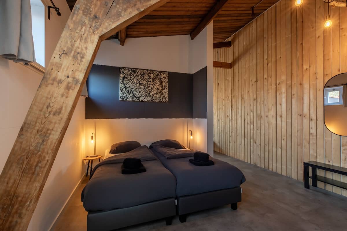A loft-style bedroom with a sloped ceiling, two beds, a wall-mounted abstract art piece, and a small window.