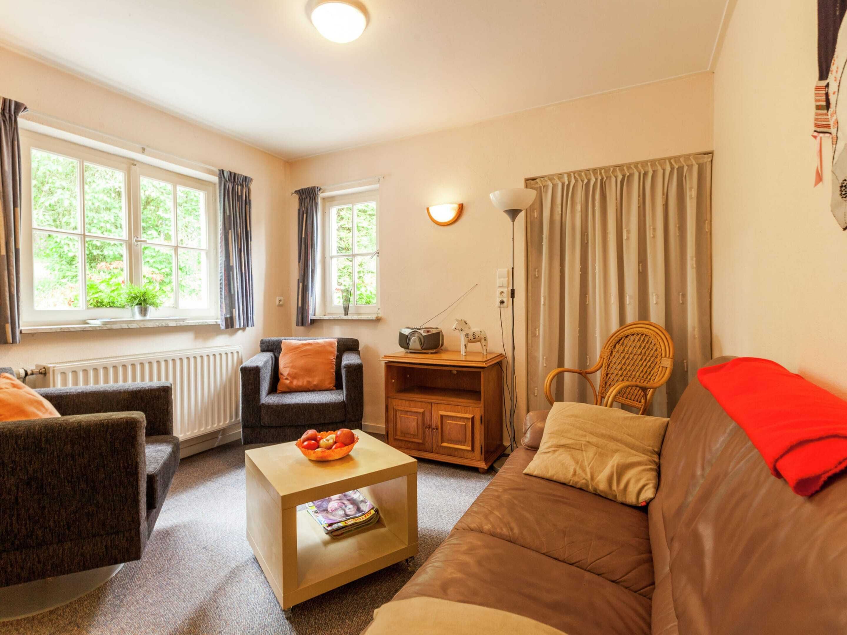A cozy living room with a brown sofa, a dark armchair, a wooden coffee table, and a wicker chair. There are two windows with curtains, a radiator, and a lamp on the wall.