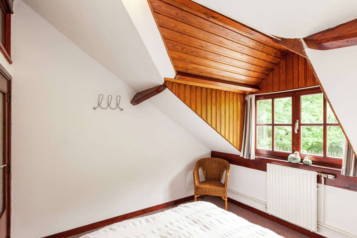 A bedroom with a sloped ceiling, a wooden chair, a radiator, and a window with curtains.