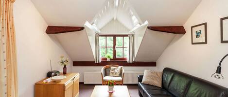 A cozy attic room with a sloped ceiling, a wooden desk, a black leather sofa, and a dining table.