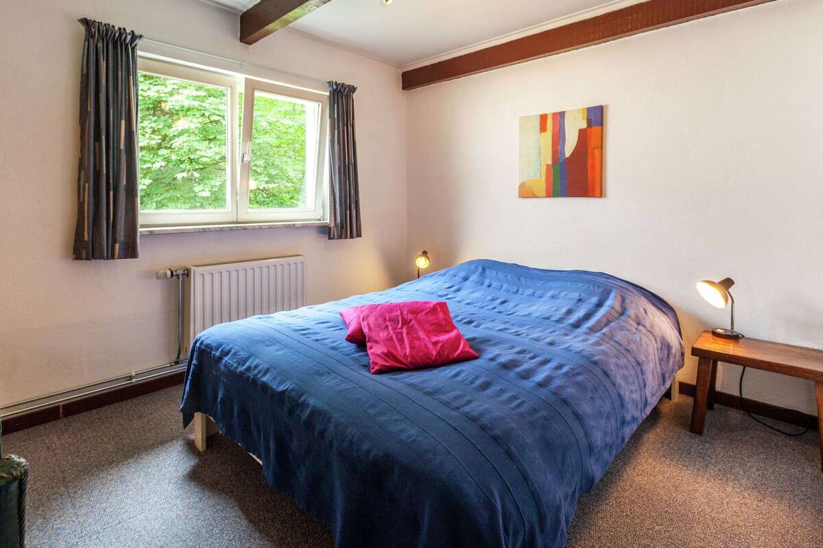 A bedroom with a large bed, a window with curtains, a radiator, a bedside table with a lamp, and a colorful abstract painting on the wall.