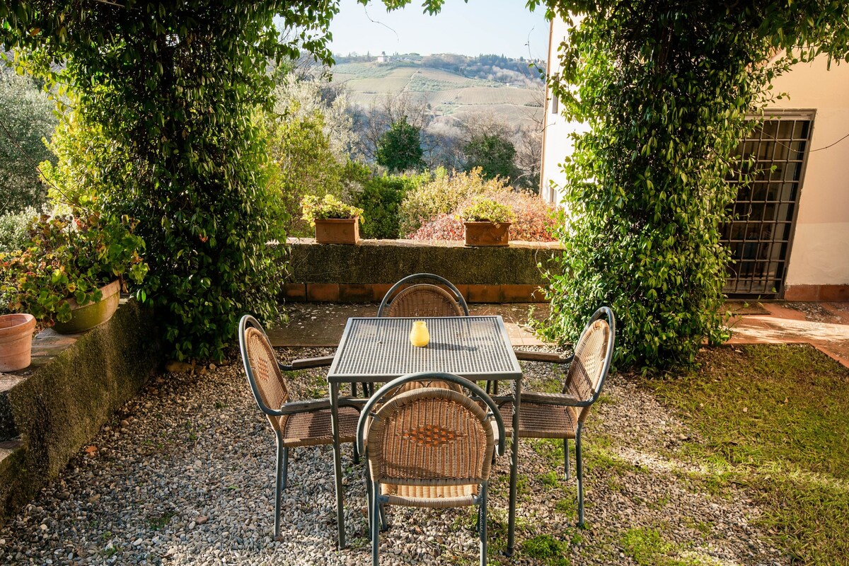 An outdoor seating area with a table and chairs, surrounded by greenery and a view of rolling hills.