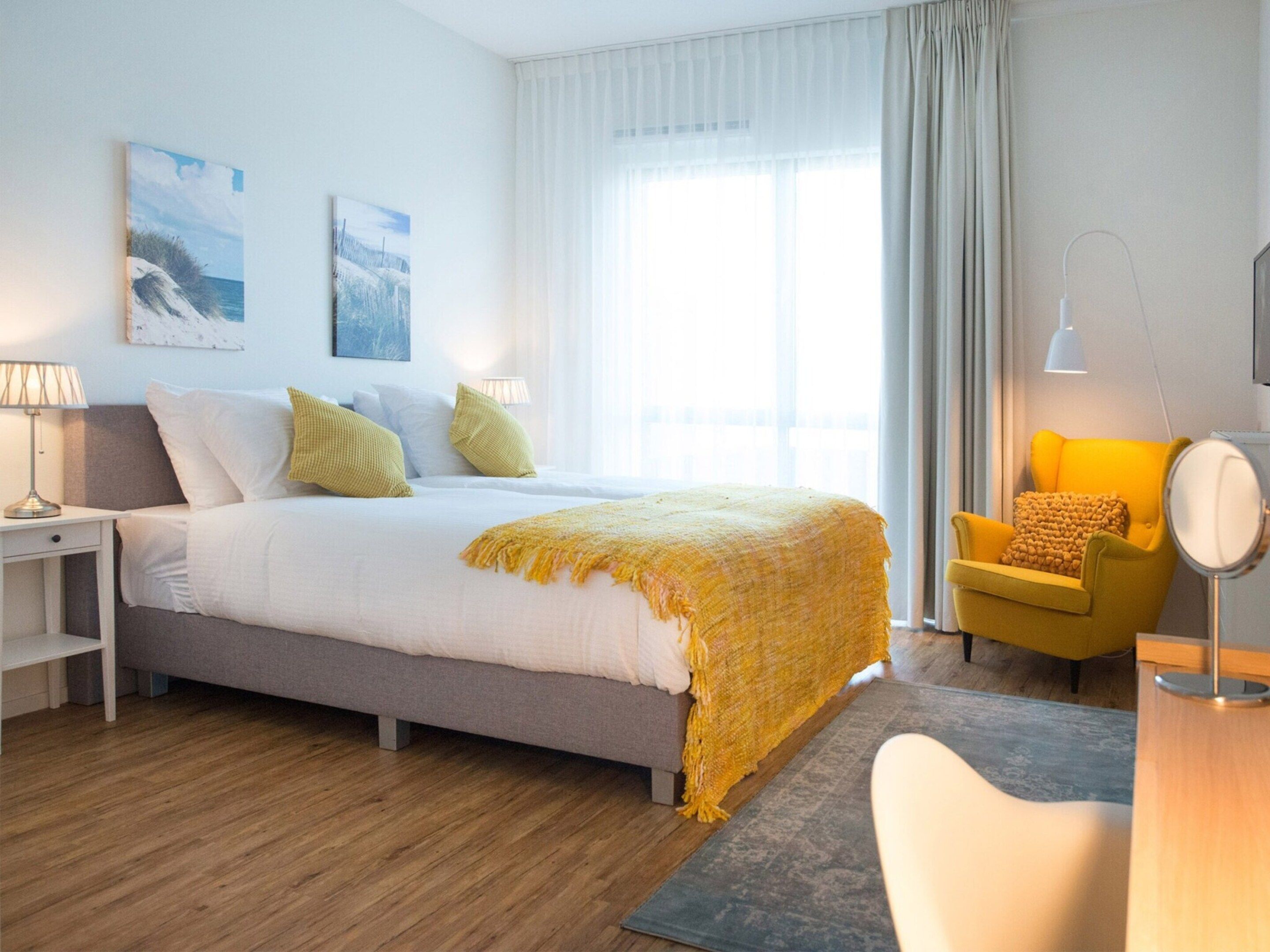 Een hotelkamer met een bed, nachtkastjes, een stoel, een bureau en een groot raam met gordijnen.