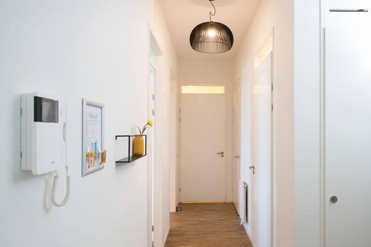 A hallway with a wooden floor, a white wall-mounted phone, a framed picture, a small shelf with a vase, and a door at the end.