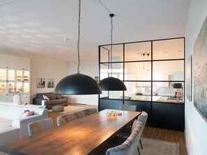A modern living room with a wooden dining table, grey sofas, and a kitchen visible through a glass partition. - Appealing Apartment in Den Haag With Balcony, Terrace (The Hague)