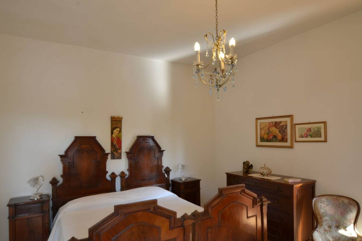Um quarto com cama de madeira, um candelabro, cômoda de madeira, uma cadeira e duas imagens emolduradas na parede.