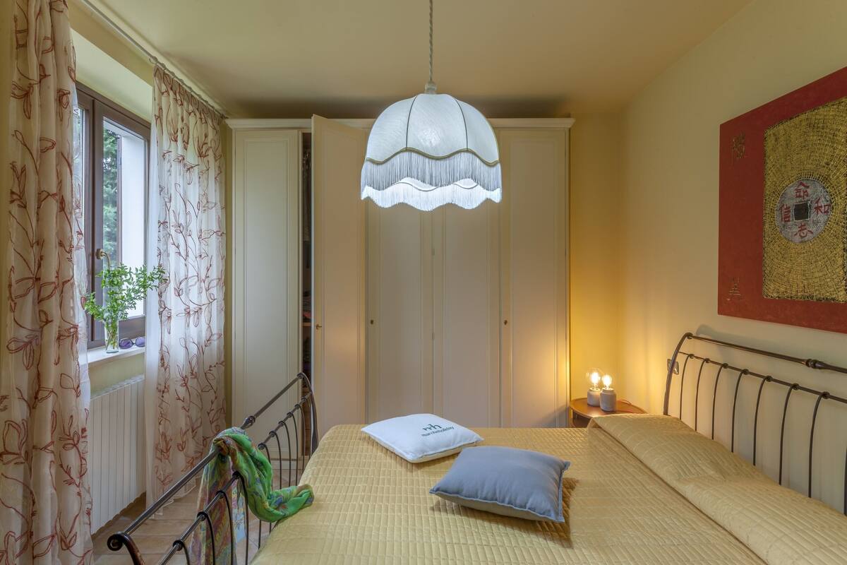 Ett sovrum med en säng, en hängande lampa, ett nattduksbord med ett ljus och ett fönster med gardiner.