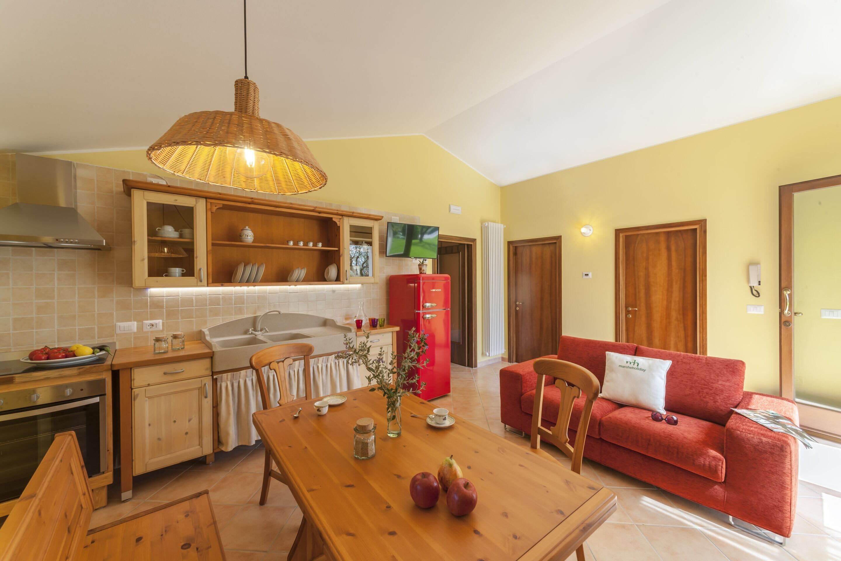 A compact kitchen with wooden cabinets, a dining table with apples, a red refrigerator, and a red sofa in a yellow-walled living area.