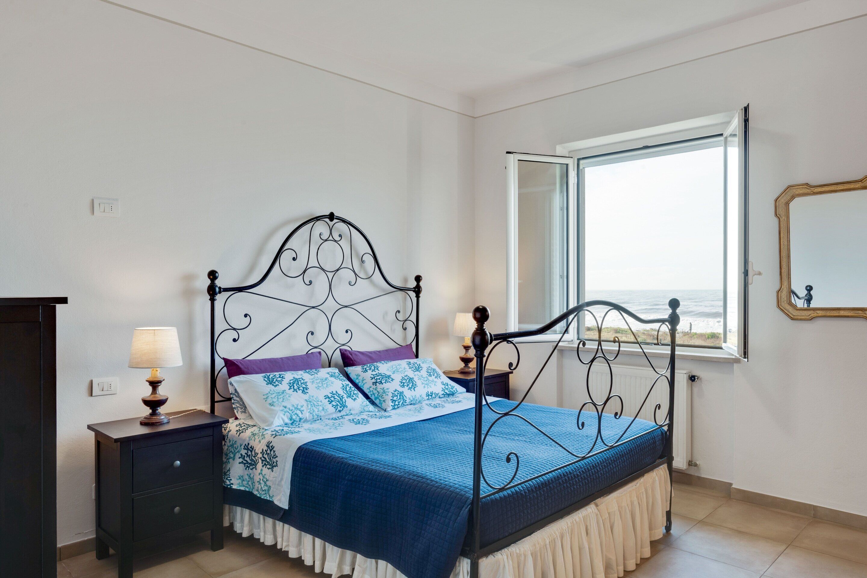 A bedroom with a bed, a nightstand, a lamp, a window with a view, and a mirror on the wall.
