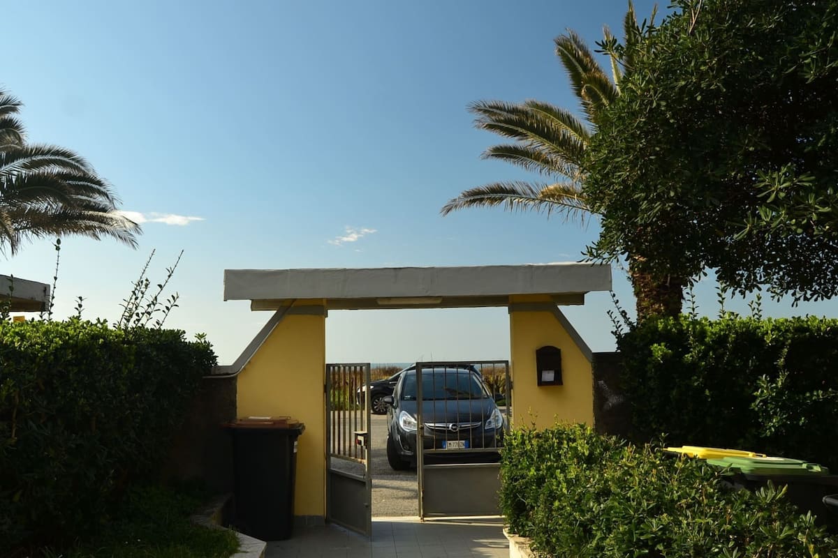 Una entrada protegida por un portón con un automóvil estacionado en su interior, rodeada de follaje y un cielo despejado.