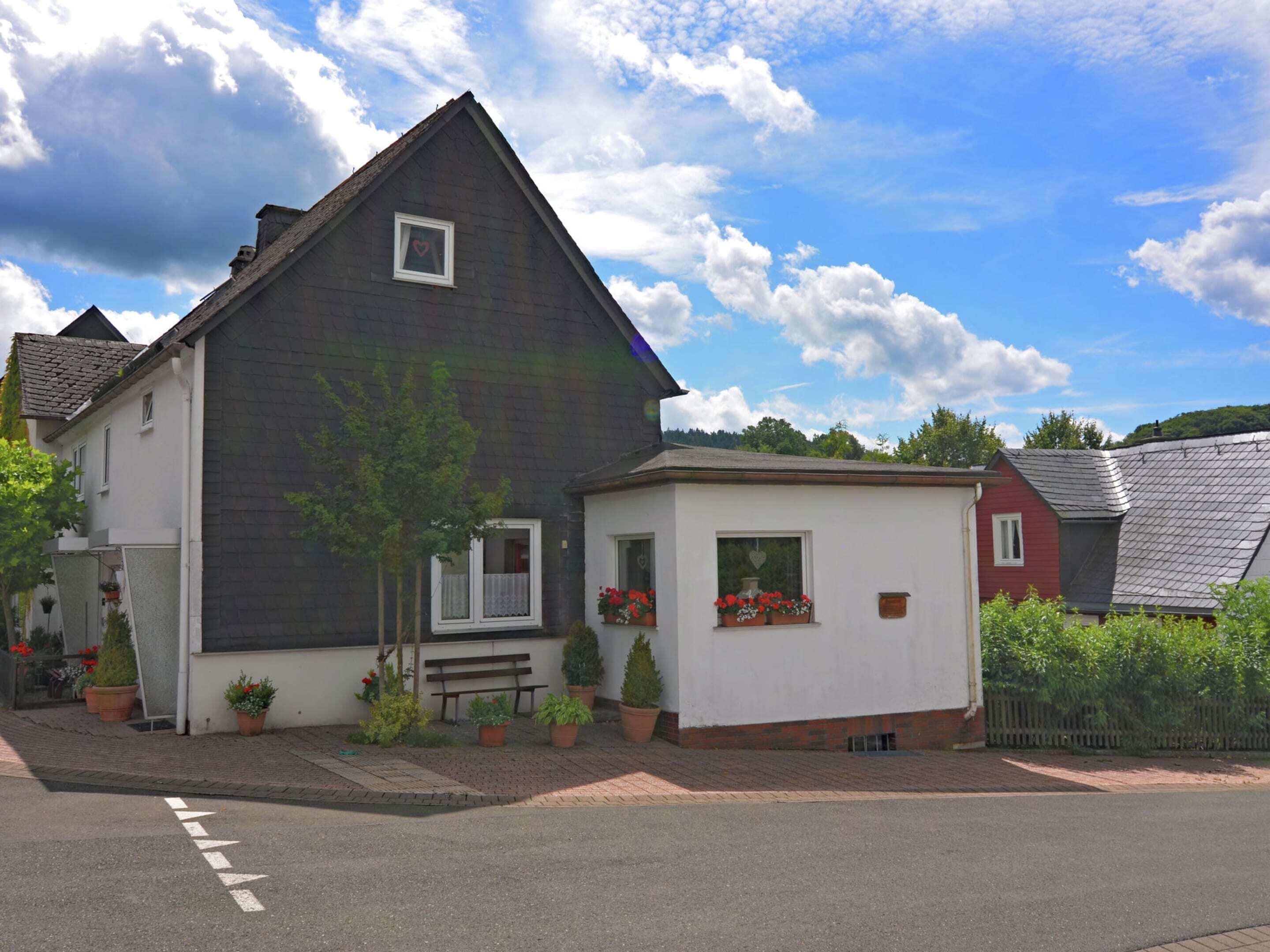 A two-story house with a dark roof, white walls, and a small attached garage. There are potted plants and a bench outside.