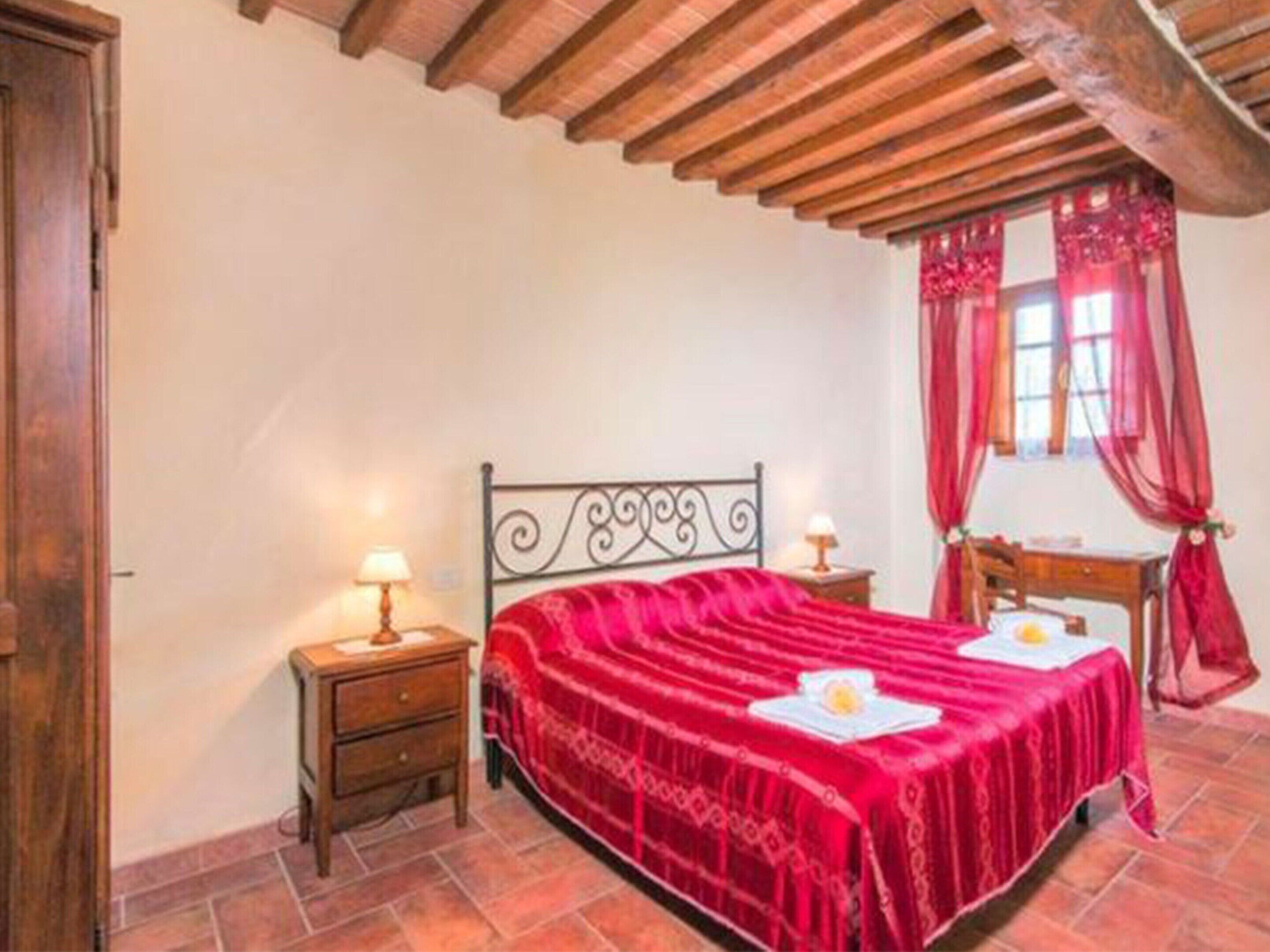 Une chambre avec un plafond en bois, un cadre de lit en métal, une couvre-lit rouge, une table de chevet avec une lampe et une fenêtre avec des rideaux rouges.