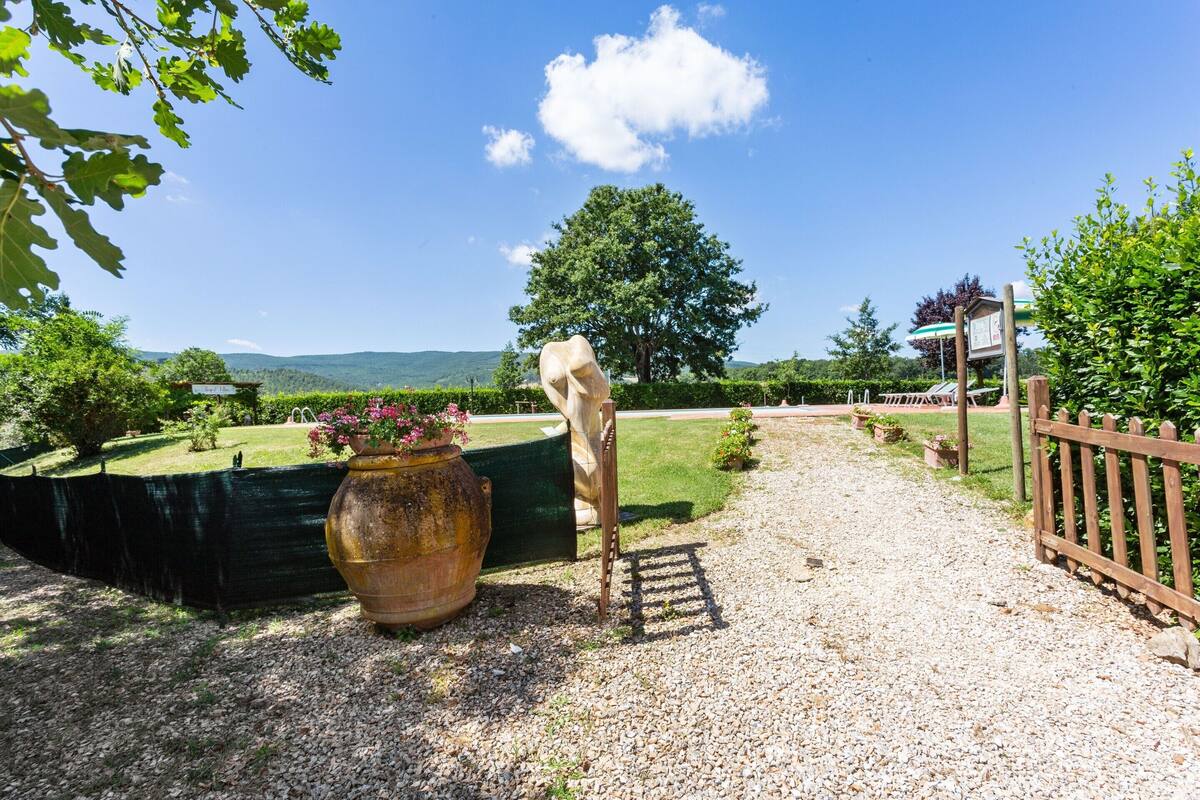 Un chemin de gravier menant à un portail en bois, un grand pot rempli de fleurs et un arbre dans un espace aménagé.