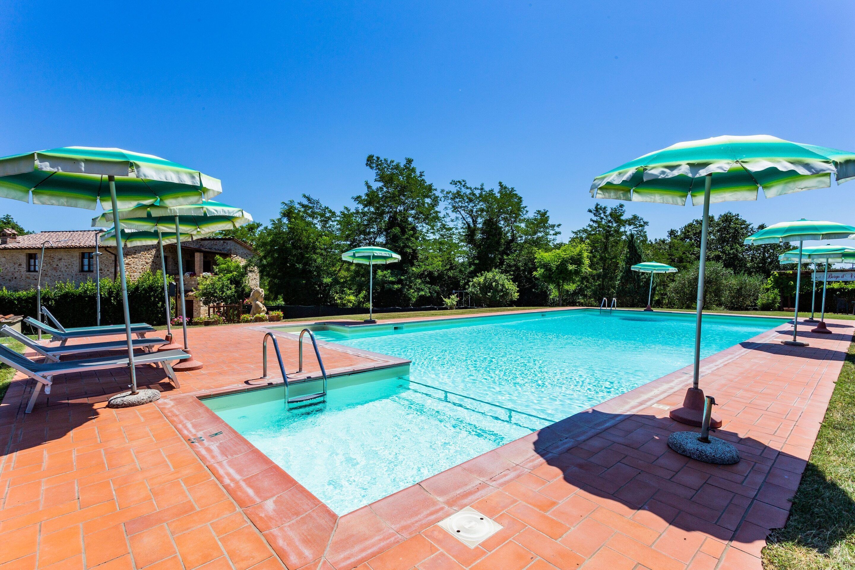 A swimming pool with a tiled deck, surrounded by green umbrellas, and a stone building in the background.