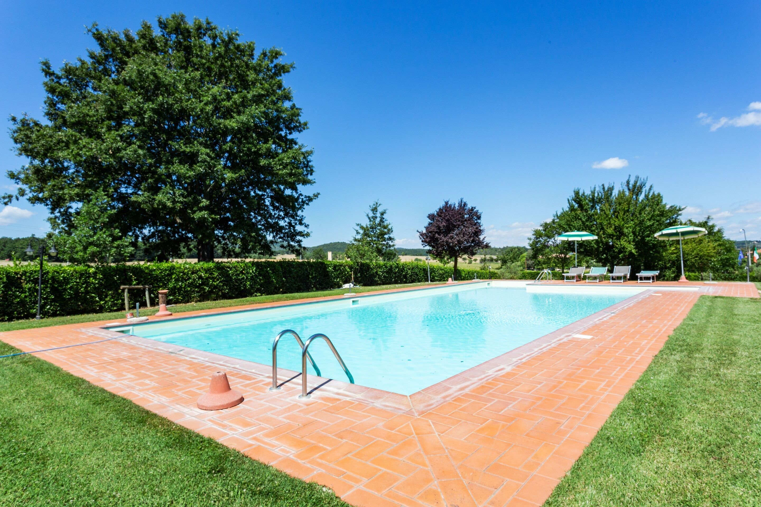 Une piscine rectangulaire, bordée d’une terrasse carrelée et entourée de pelouse et d’arbres.