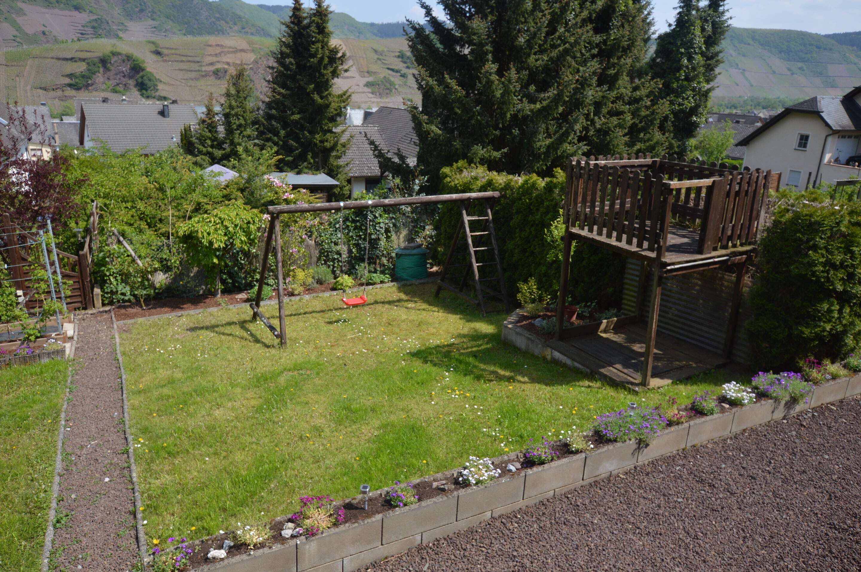 Ein Garten mit einer Holz-Spielanlage, einem Blumenbeet und Blick auf Häuser und Hügel im Hintergrund.