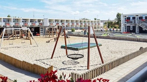 Un terrain de jeux équipé de balançoires et d’un trampoline, entouré d’une zone sablonneuse et bordé d’un bâtiment doté de plusieurs balcons.