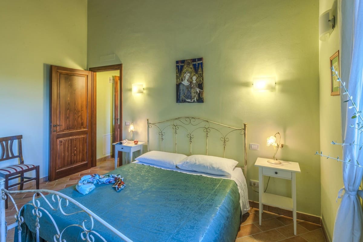 Ein Schlafzimmer mit einem Bett, zwei Nachttischen, einem Hocker, einem Stuhl, einer Holztür und einem an der Wand befestigten Bild.