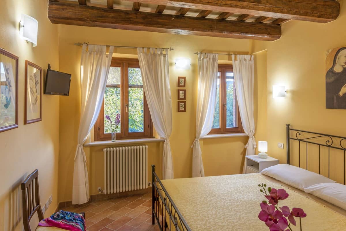 Ein Schlafzimmer mit einem Bett, zwei Fenstern mit Vorhängen, einem Heizkörper, einem Fernseher und einem Bild an der Wand.