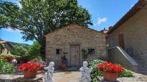 A stone building with a wooden door, potted red flowers, and two stone statues. - Detached Villa for 6-pers. With Outdoor Swimming Pool (Loro Ciuffenna)