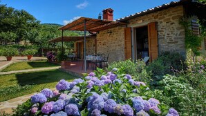 A stone building with a tiled roof, a covered patio with outdoor seating, and a garden with hydrangea flowers. - Detached Villa for 6-pers. With Outdoor Swimming Pool (Loro Ciuffenna)