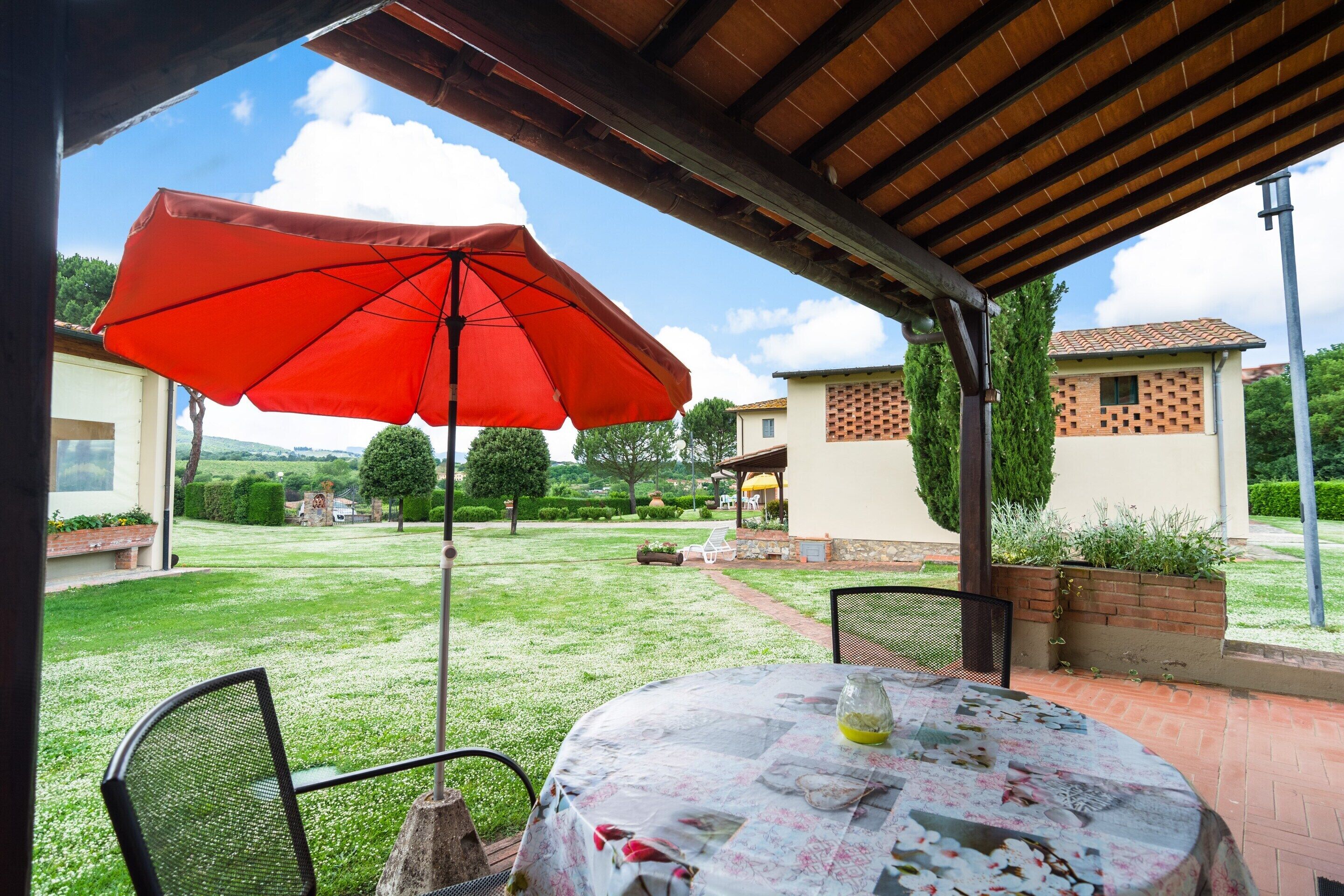 Um pátio externo com mesa e cadeiras, um guarda-sol vermelho e vista para um jardim e edifícios ao fundo.