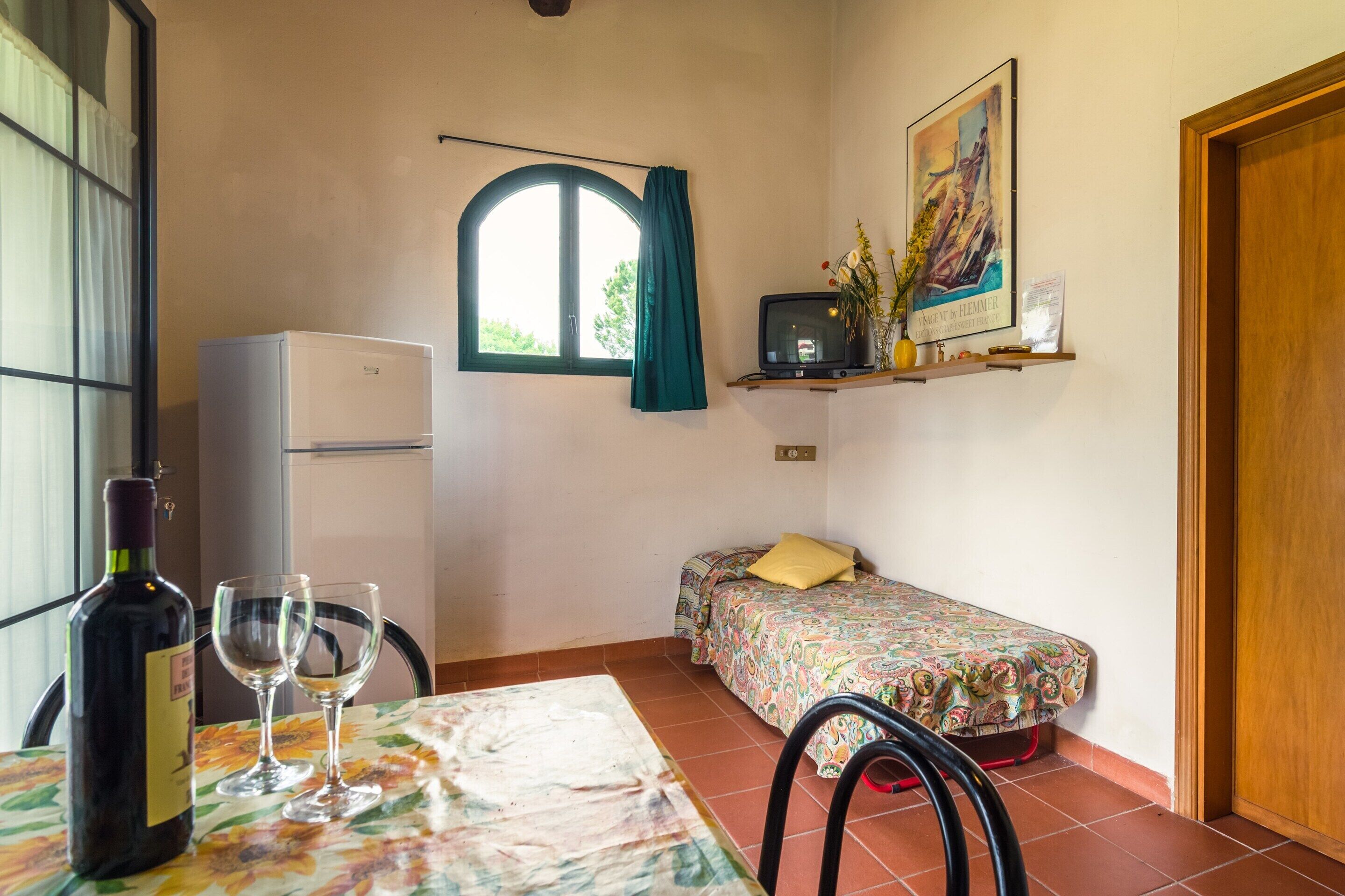A small room with a bed, a table with a bottle and glasses, a TV, and a window with curtains.