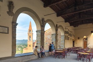 Un café en plein air avec des arches en pierre, une tour de l’horloge en arrière-plan, et des tables et des chaises disposées pour accueillir les clients.