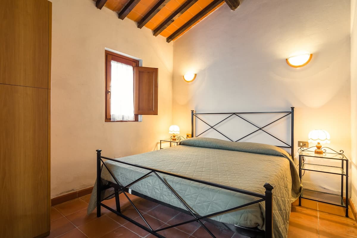 Een slaapkamer met een bed, houten balken in het plafond, twee bedlampjes en een raam met houten luik.