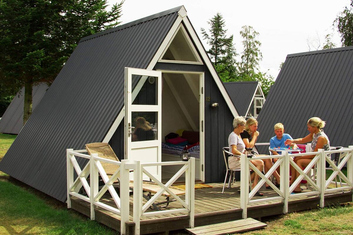 Une petite structure à toit triangulaire, dotée d’une terrasse en bois et de rampes blanches, entourée d’herbe et d’arbres.
