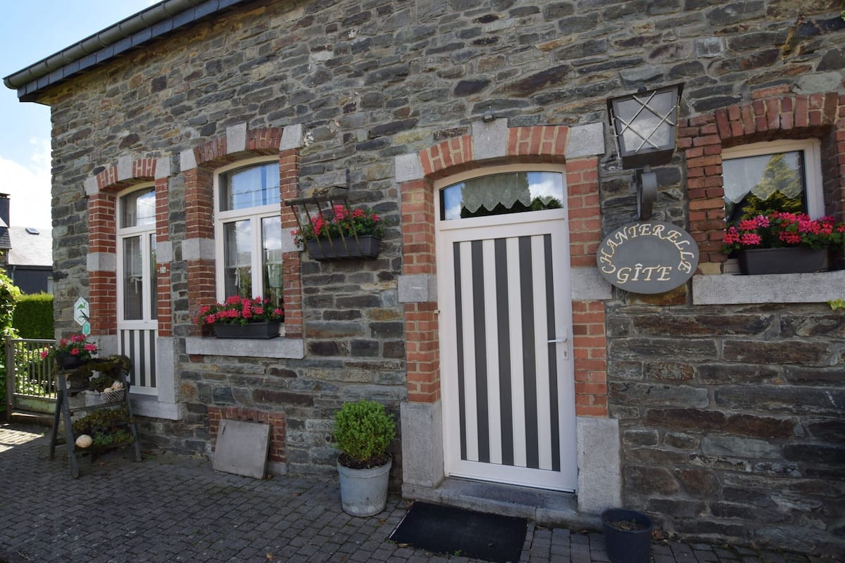 A stone building with a white door and windows, a sign reading "HANSERELLE Gîte", and potted plants on the windowsills.