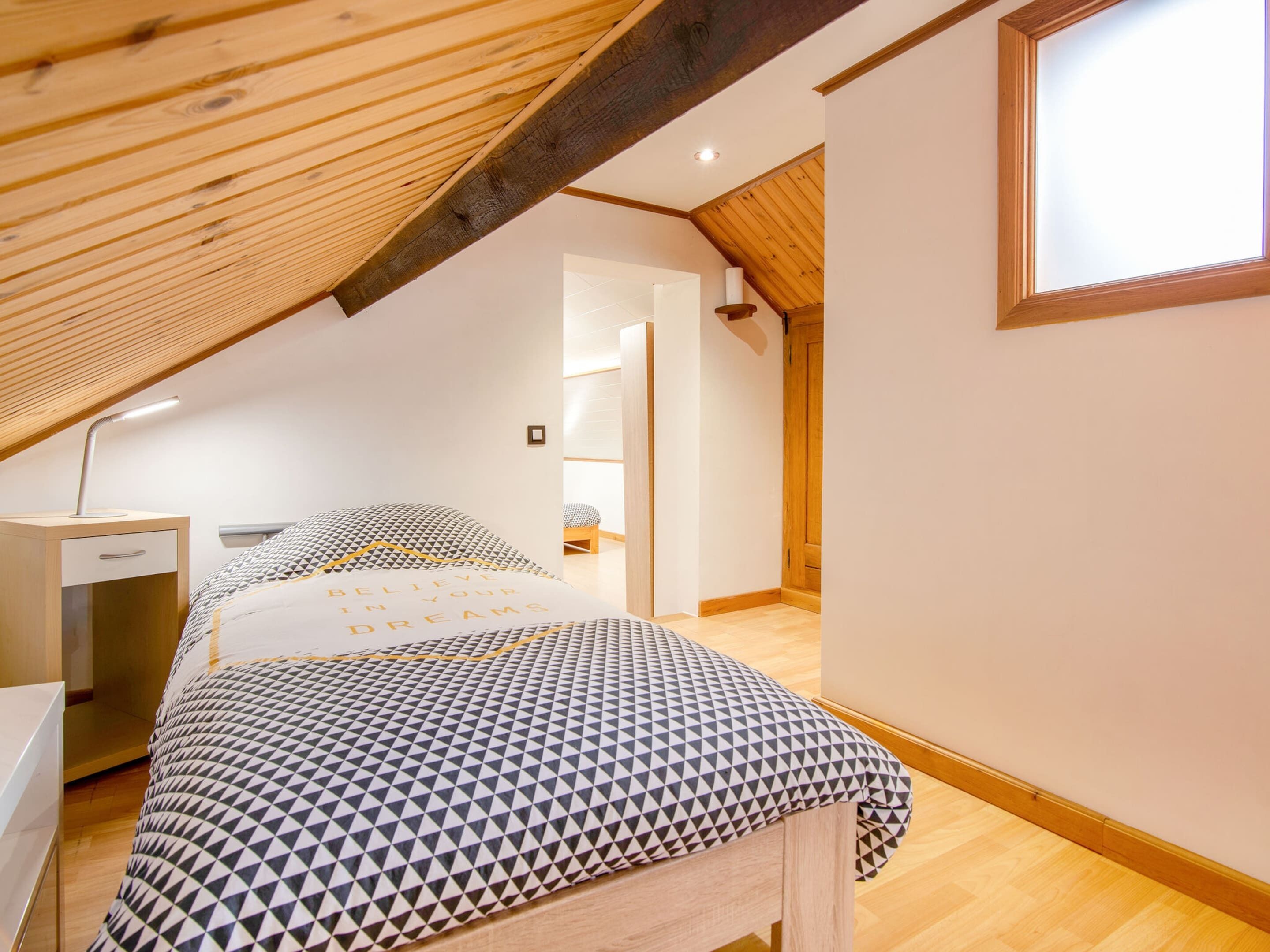 A bedroom with a sloped ceiling, a bed with a geometric-patterned bedspread, a bedside table, and a window.