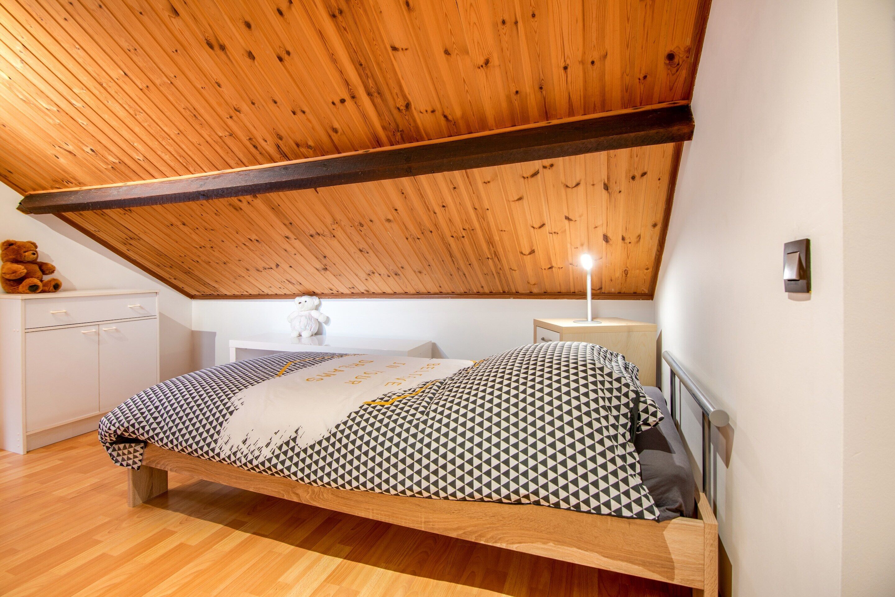 Un dormitorio con techo inclinado, una cama con ropa de cama estampada, una mesita de noche con una lámpara y un oso de peluche en un aparador.