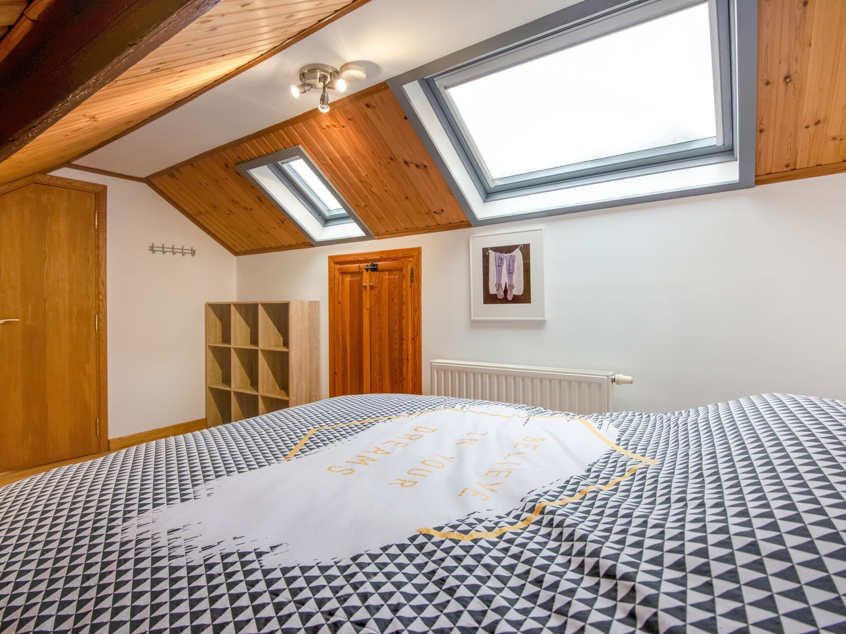 A bedroom with a skylight, a bed with patterned bedding, a wooden wardrobe, and a radiator.