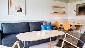 Ett modernt vardagsrum med en grå soffa, ett runt bord och trästolar. Det finns två vägghängda hyllor och en taklampa.
