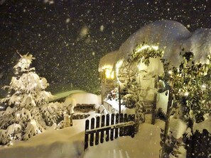Een besneeuwde buitenruimte met een houten hek, een met sneeuw bedekte boom en een gebouw met een verlicht raam.