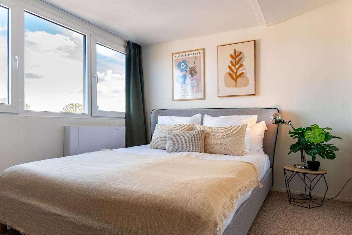 A neatly made bed with a beige blanket, a grey headboard, and multiple pillows. There is a window with a view of the sky and clouds, and a small round table with a plant and a lamp.