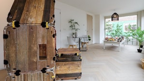 Un interior moderno con una estructura de madera que recuerda a una pila de maletas, una zona de comedor con mesa y sillas, y vistas a la vegetación a través de un ventanal.