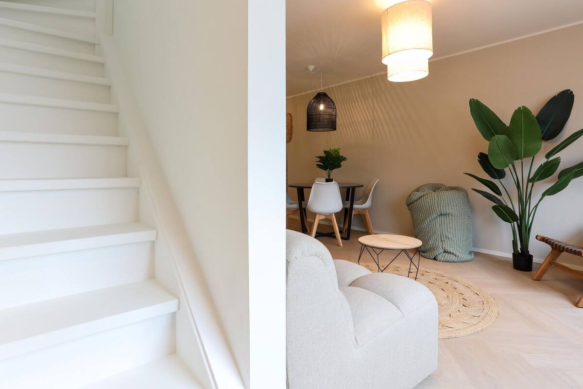 A staircase leading to a living area with a dining table, chairs, a sofa, and a large plant.