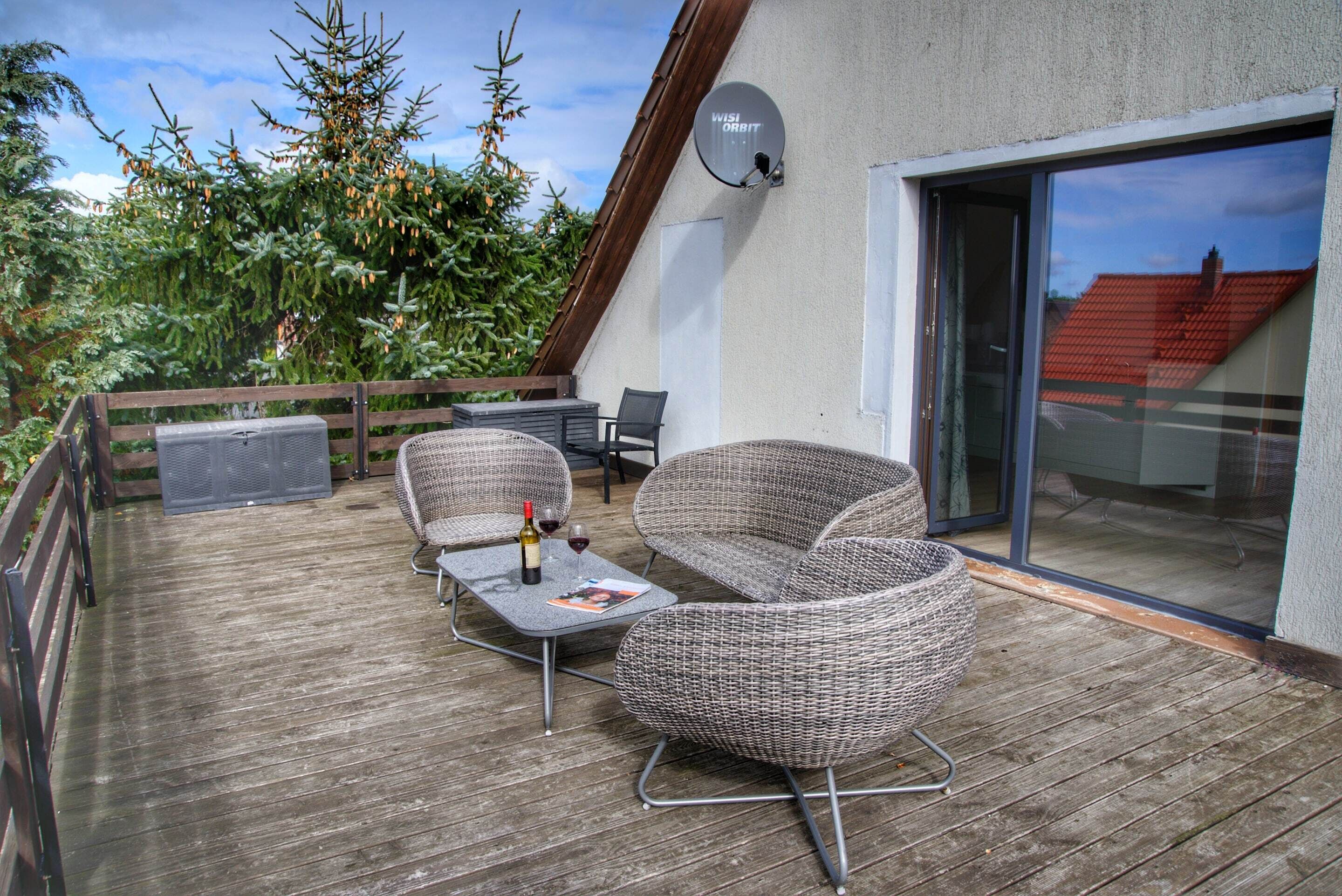 Un terrazza in legno con mobili in vimini, un'antenna parabolica e una porta vetrata che dà su un edificio con tetto rosso.