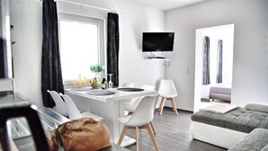 A modern living room with a sofa, a dining area with a table and chairs, a flat-screen TV mounted on the wall, and a window with curtains. - Unterkunft in Monkebude mit Eigenem Garten (Monkebude)