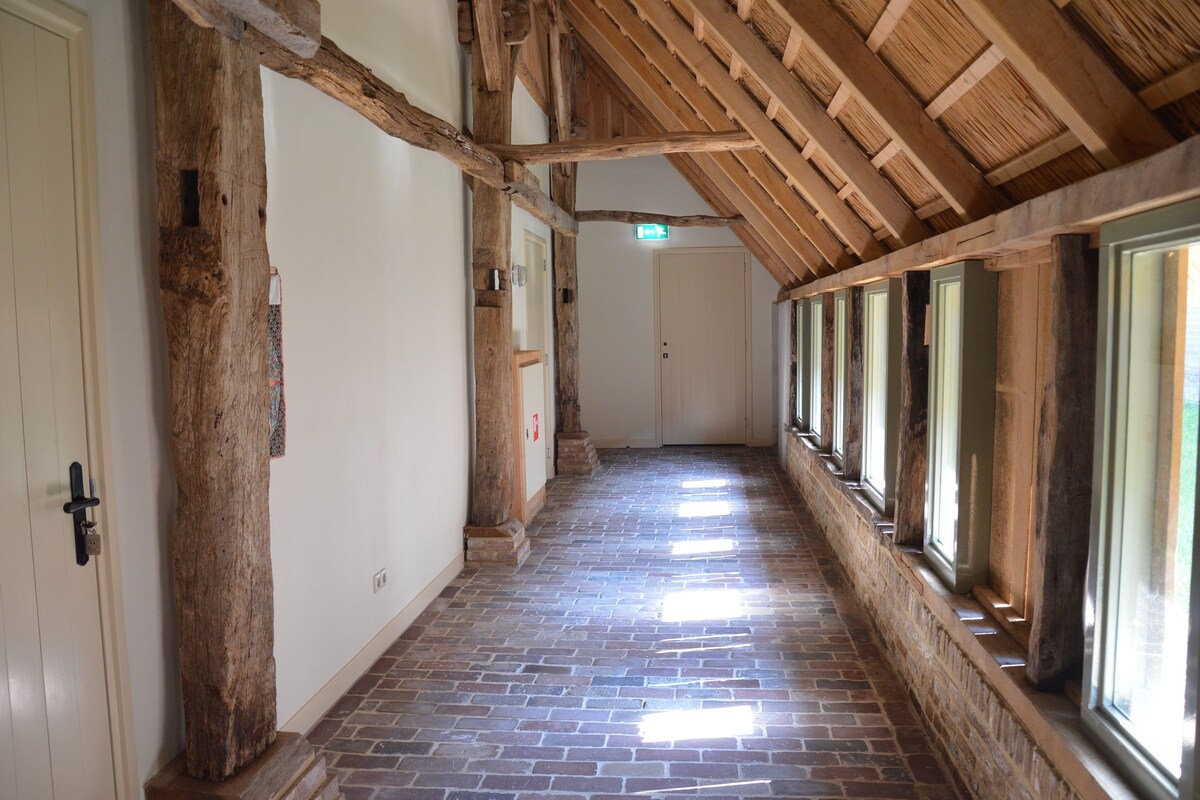 A hallway with exposed wooden beams, brick flooring, and large windows along one side.