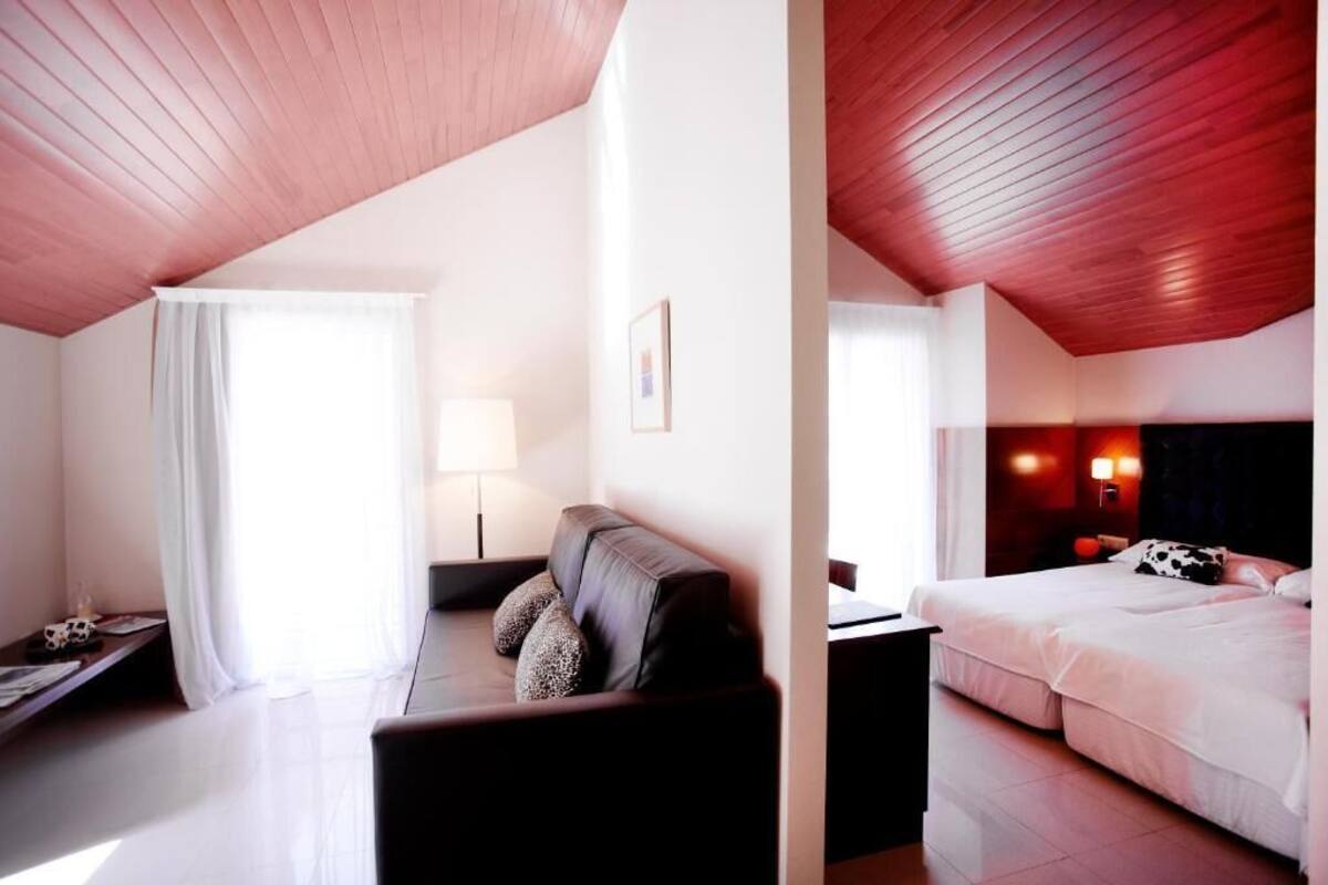 A hotel room with a wooden ceiling, a bed with white bedding, a sofa with patterned cushions, a bedside table, and a lamp.