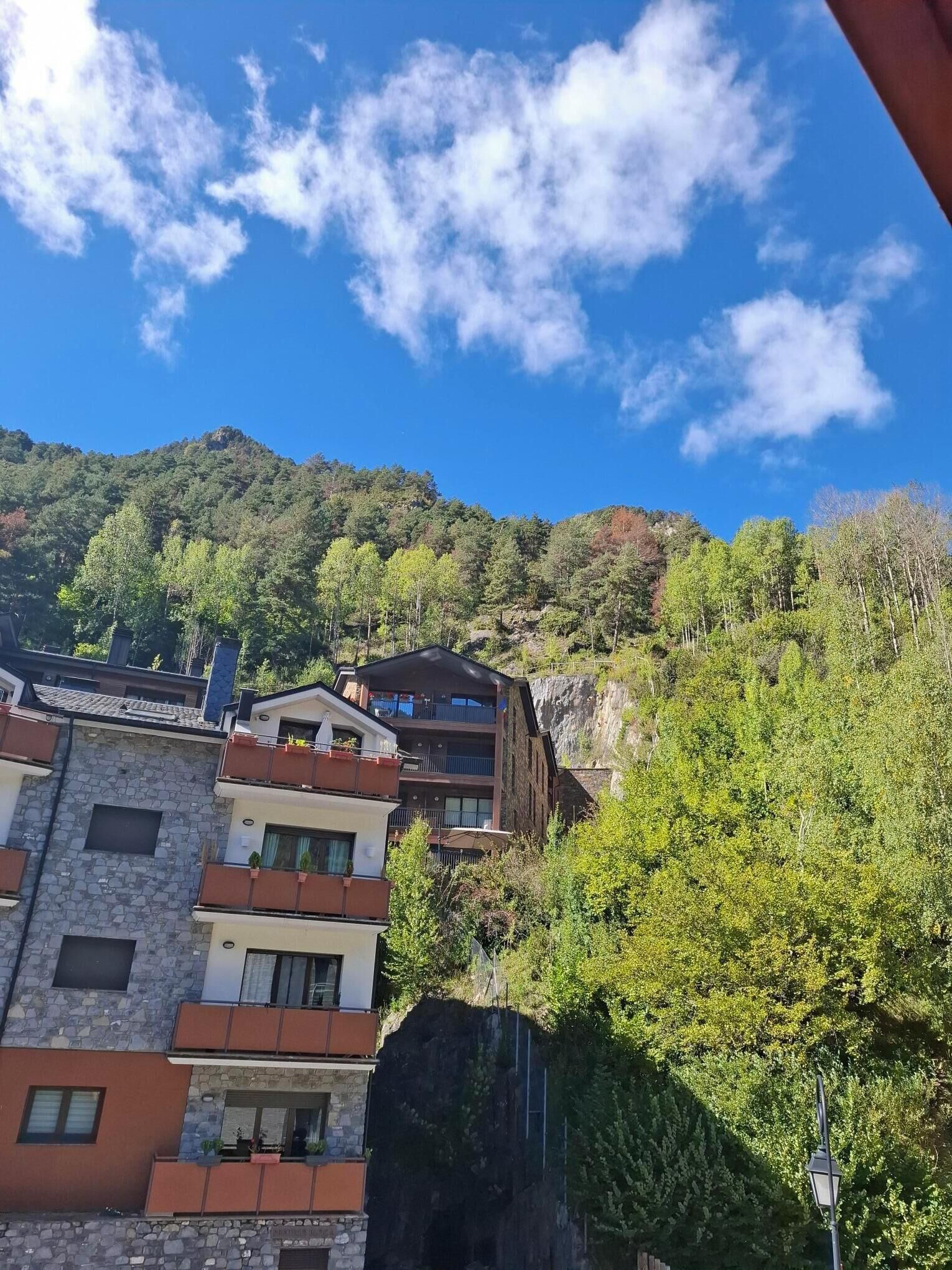 Un edificio de varios pisos con balcones, con el telón de fondo de una ladera boscosa y un cielo azul despejado.
