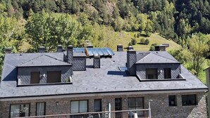 A two-story building with a stone facade and multiple chimneys. The building has a sloped roof with solar panels and a small balcony. - Hotel Mu (La Cortinada)