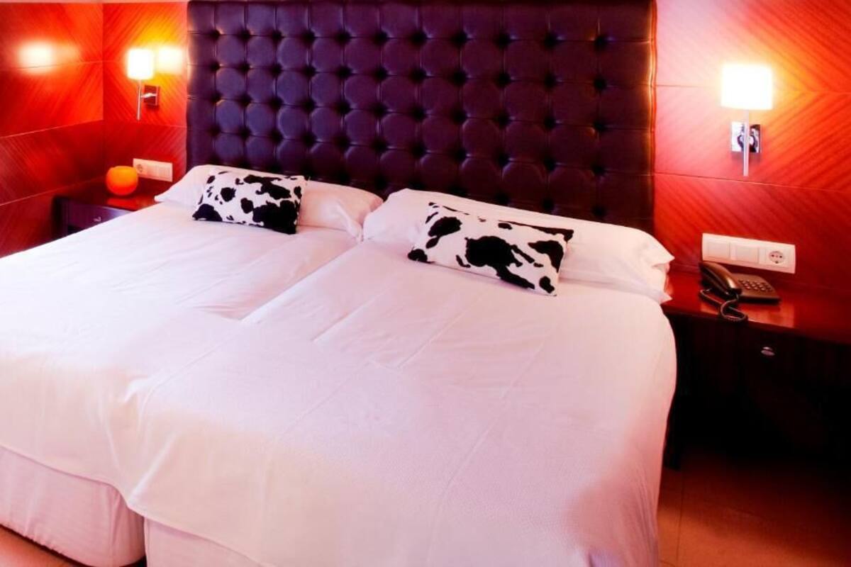 A double bed with white bedding and black and white cow-patterned pillows. The headboard is tufted and dark-colored. There are two bedside tables, one with a lamp and the other with a telephone. The walls are red with a textured finish.
