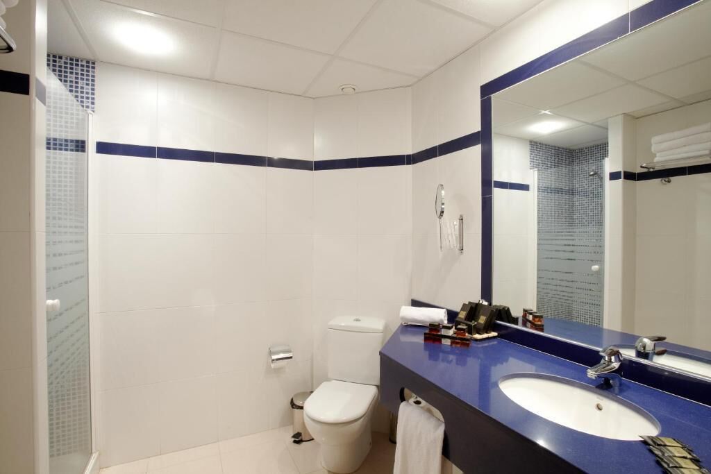 A bathroom with white tiles, a blue-tiled shower area, a white toilet, and a sink with a mirror.