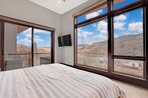 A bedroom with a large bed, a TV mounted on the wall, and a balcony with a view of the mountains. - Luxurious Contemporary Home (Silverthorne)