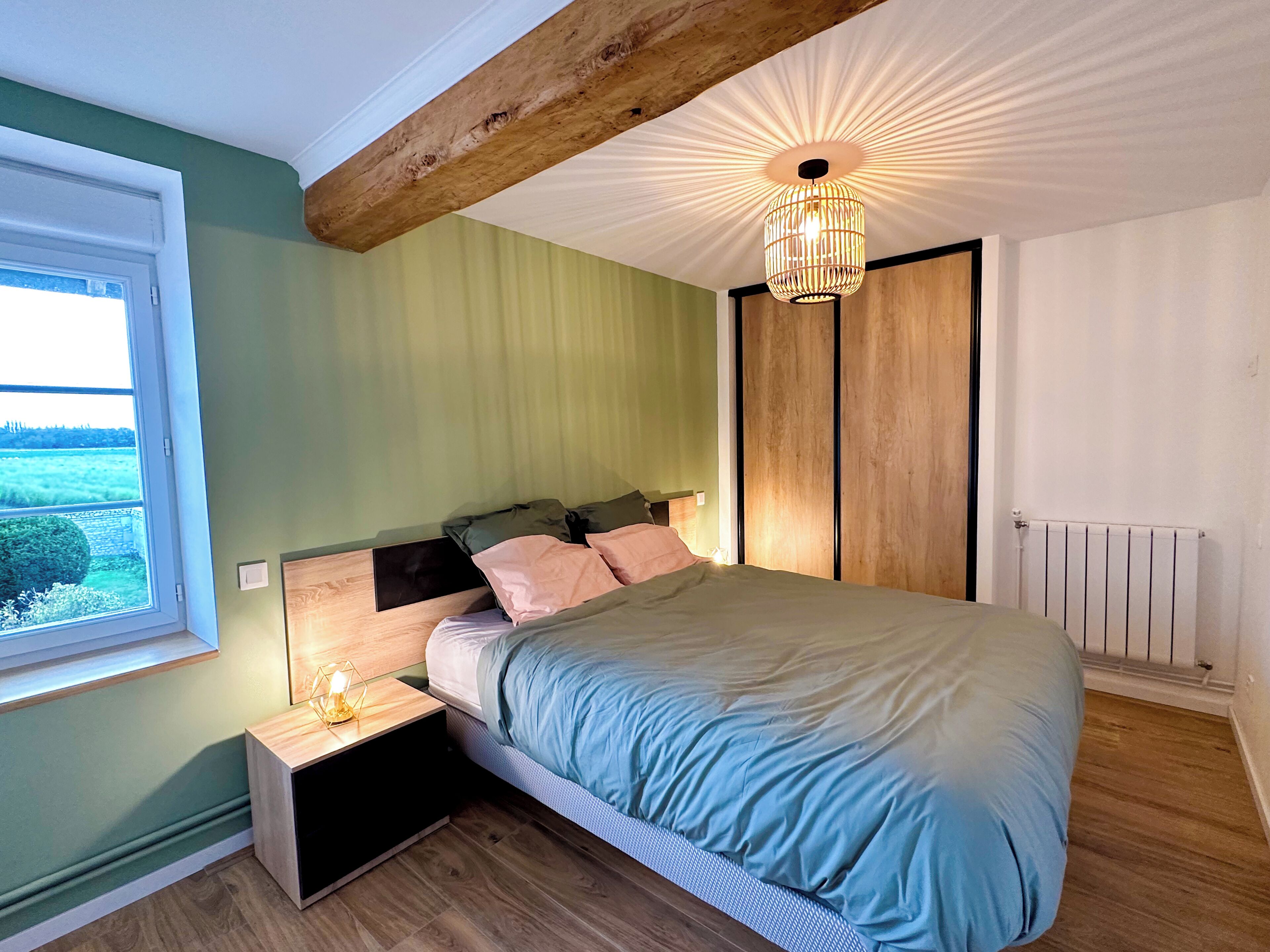 Ein Schlafzimmer mit einem Bett, einem Nachttisch, einem Fenster, einem Holzbalken an der Decke und einer Heizung.