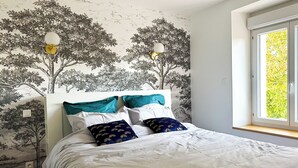 Een slaapkamer met een bed, witte beddengoed, kussens in teal-kleur en een wanddecoratie van bomen.